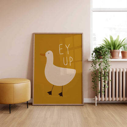 A Dandy Prints framed large artwork of a hand illustrated duck on a mustard yellow background with the Yorkshire slang 'EY UP' written above the duck. In a living room by a windowsill with plants and a yellow footstool.