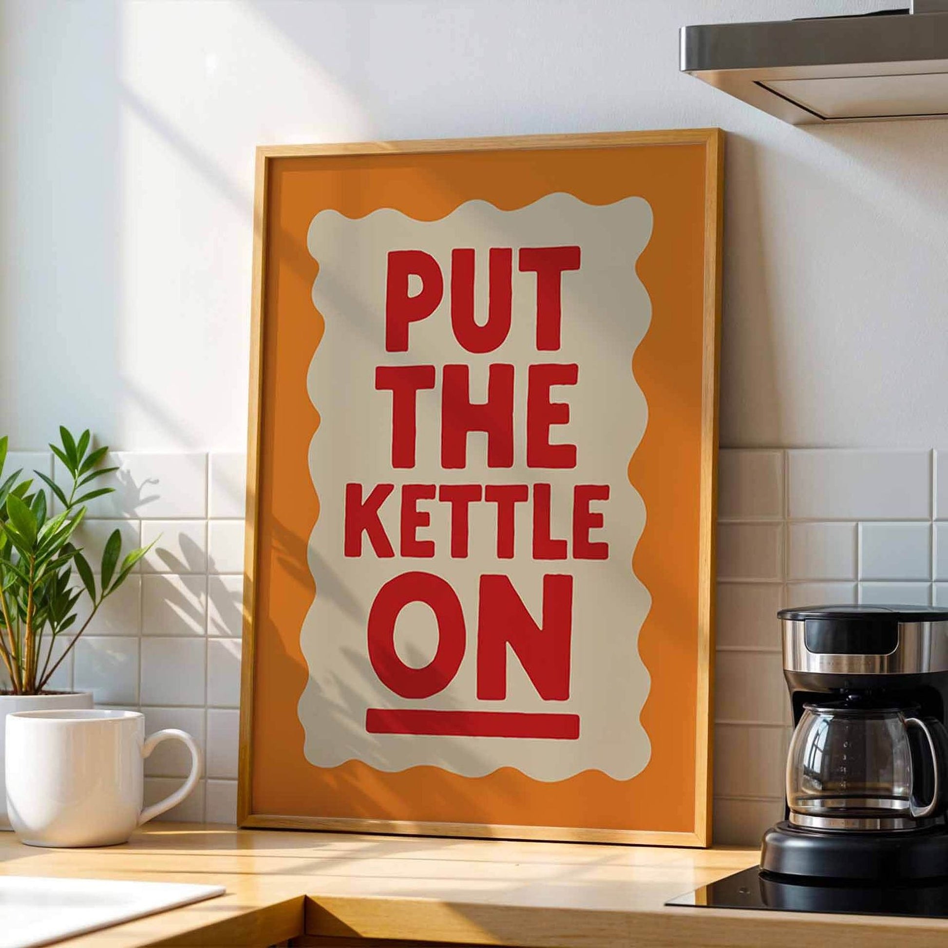 a framed poster with the text "PUT THE KETTLE ON" in bold red letters, placed on a kitchen counter next to a coffee maker.